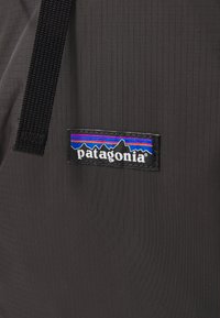 Crna tkanina s teksturom mreže s oznakom logotipa Patagonia koja prikazuje višebojni planinski grafički element i tekst. Povez crne boje je pričvršćen.