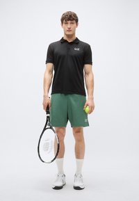 Juodas polo marškinėlis ir žalsvi šortai, laiko teniso raketę ir kamuoliuką. Audinys su tekstūra, sportiškas siluetas. Baltos sportinės bateliai ir vidutinio ilgio kojinės.