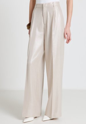Pantalon beige taille haute à jambes larges avec plis, porté avec des chaussures blanches à bout pointu et un bracelet en bois au poignet gauche.