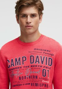 Roter Strickpullover mit marineblauem Text, Rundhalsausschnitt und bedruckten Grafiken, darunter "CAMP DAVID" und geografische Verweise.