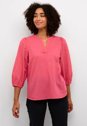 Rosa, kurzärmelige Bluse mit V-Ausschnitt und gerafften Puffärmeln; mit kleinen, erhabenen Punkten versehen. Kombiniert mit dunklen Hosen.