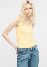Jeune femme aux cheveux blonds ondulés mi-longs, portant un haut jaune sans manches et un jean bleu, se tenant debout avec une main sur la hanche.