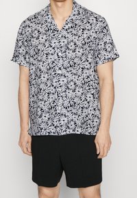 Hombre vistiendo una camisa de botones de manga corta, blanca y negra con estampado floral, combinada con pantalones cortos negros lisos, de pie frente a un fondo neutro.
