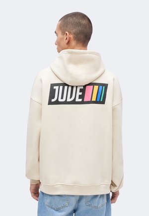 Homme portant un sweat à capuche crème avec le texte "JUVE" et des rayures verticales roses, jaunes, bleues et vertes sur un rectangle noir dans le dos, jean bleu.