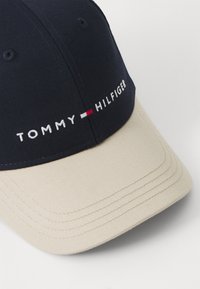 Marineblå baseball cap med en lys brun skygge, prydet med broderet "TOMMY HILFIGER" logo i hvid, rød og blå. Glat stof med struktureret design.