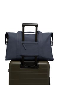 Sac de sport bleu marine avec une poche zippée et deux poignées, reposant sur une valise vert olive foncé avec une poignée télescopique.