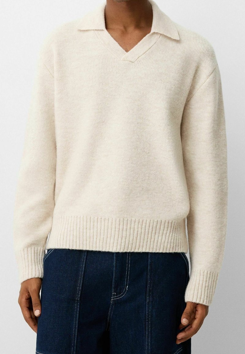 Homme portant un pull en maille couleur crème avec un col en V et un pantalon bleu foncé avec des coutures blanches visibles.