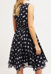 Marineblaues Polka-Dot-Kleid mit figurbetontem Oberteil, fließendem Rock und ärmellosem Design mit taillierter Taille. Der Stoff ist glatt und durchsichtig.