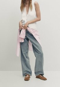 Gilet bianco a coste con scollatura a V profonda, abbinato a jeans a gamba larga blu chiaro. Camicia rosa chiaro annodata in vita. Scarpe marroni slip-on.