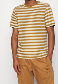 Stribet t-shirt i sennepsgul og lys blå. Korte ærmer, rund hals, blødt bomuldsstof. Sammen med beige bukser med en lomme.