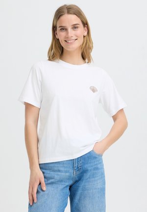 Jeune femme souriante, portant un t-shirt blanc avec un petit motif coquillage marron sur la poitrine, et un jean bleu clair, debout avec une main dans la poche.