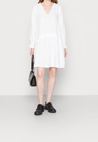 Vestido blanco de manga larga con escote en V y cintura con volantes. Combinado con zapatos de plataforma negros y un pequeño bolso negro.