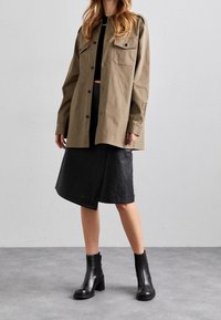 Khaki oversized skjorte med to brystlommer, parret med en sort læderskirt med en vinkelret kant. Sorte ankelstøvler fuldender looket.