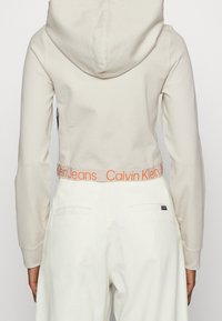 Widok z tyłu osoby w beżowej krótkiej bluzie z kapturem i białych spodniach o wysokim stanie, z napisem "Calvin Klein Jeans" na pasku.