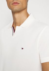 Polo bianco con colletto, caratterizzato da una striscia colorata sul collo e un piccolo logo sul petto. Texture liscia, maniche corte.