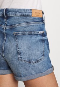 Pantalones cortos de mezclilla azul con dobladillo doblado, que cuentan con una etiqueta de cuero en la cintura y dos bolsillos traseros con detalles de costura.