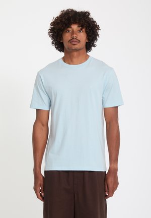 Uomo con capelli ricci che indossa una maglietta azzurro chiaro a maniche corte e pantaloni marrone scuro, in piedi davanti a uno sfondo bianco semplice.