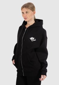Sweat à zip noir en tissu doux, avec une poche kangourou et un graphique blanc sur la zone gauche de la poitrine.
