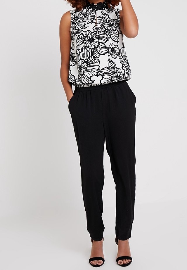 Femme portant un chemisier sans manches à motif floral noir et blanc, un pantalon noir taille haute et des talons ouverts noirs, debout avec une main dans la poche.