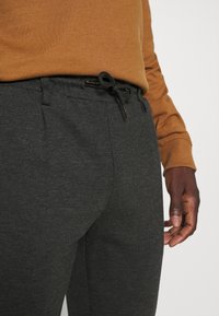 Pantalons de survêtement gris foncé avec un cordon de serrage à la taille, présentant une texture douce et des poches latérales. Portés avec une chemise à manches longues marron.