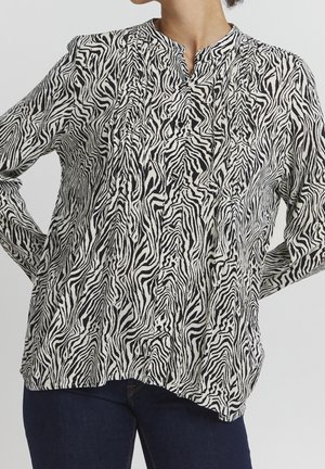 Schwarz-weiße Bluse mit Zebramuster, Mandarin-Kragen, langen Ärmeln, gerafftem Vorderteil und entspannter Passform, kombiniert mit dunklen Jeans.