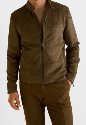 Homme portant une veste en daim marron à fermeture éclair sur une chemise marron et un pantalon marron assorti, une main dans la poche, sur fond blanc.
