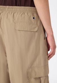 Beżowe spodenki cargo z elastycznym paskiem i tylną kieszenią z klapką noszone przez osobę z widoczną prawą ręką i ciemną górą.