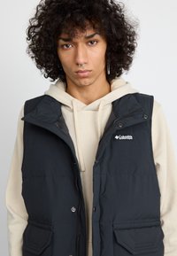 Colete isolante preto com botões de pressão, fecho éclair, dois bolsos frontais e logotipo da Columbia, sobreposto a um hoodie creme. Textura suave.