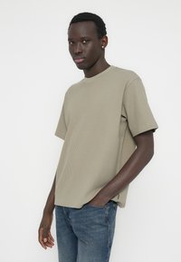 Λιγν πράσινο ριμπ μπλουζάκι με χαλαρή εφαρμογή, στρογγυλή λαιμόκοψη και κοντά μανίκια. Φοριέται με μπλε τζιν, παρουσιάζοντας μια ομαλή υφή.