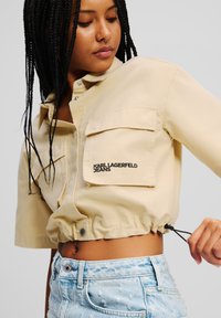 Giacca cropped beige con due tasche frontali, orlo con cordoncino e logo "KARL LAGERFELD JEANS". Indossata con pantaloni shorts in denim chiaro strappati.