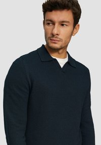 Marineblauw gebreid trui met een gestructureerd patroon, voorzien van een klassieke kraag en lange mouwen, gedragen over een wit shirt.