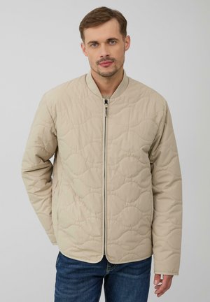 Beige gewatteerde jas met een staande kraag, rits aan de voorkant en een getextureerd patroon. Draagt over een blauwe spijkerbroek, met een losse pasvorm en een gladde stof.