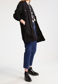 Czarna, długorękawowa kurtka bombowca z ściągaczami, zapinana na zamek, z bocznymi kieszeniami, noszona na wzorzystą koszulę, w zestawieniu z przyciętymi niebieskimi dżinsami i błyszczącymi botkami za kostkę.