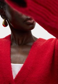 Cardigan rosso in lana con scollatura a V profonda, caratterizzato da un motivo di maglia testurizzato. Accompagnato da orecchini dorati e rossi, che evidenziano contrasti di design audaci.