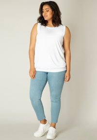 Witte mouwloze top met een ronde hals, gecombineerd met lichtblauwe skinny jeans en witte sneakers, staand tegen een neutrale achtergrond.