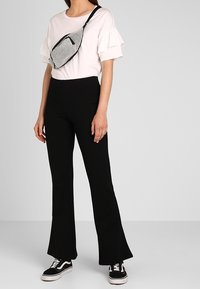 T-shirt bianco con maniche a volants, abbinato a pantaloni neri a campana e sneakers nere. Una borsa a tracolla in rete argentata con zip è indossata sul corpo.