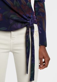Nudo hecho a mano en la parte de la cintura de una blusa de manga larga azul oscura con estampado floral, combinada con pantalones blancos que tienen un pequeño diseño de corazón dorado cerca del bolsillo.
