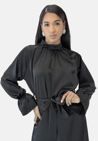 Zwarte satijnen jurk met een hoge halslijn, pofmouwen met geplooide manchetten en een taillekoorddetail. Gladde textuur met subtiele glans.