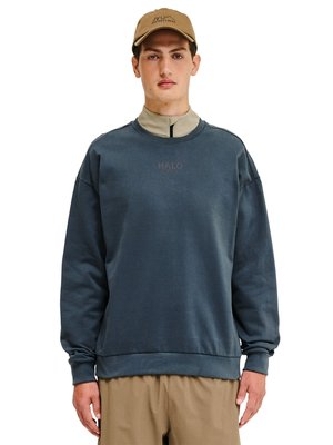 Junger Mann trägt einen blauen, übergroßen Sweatshirt mit dem Schriftzug „HALO“, beige Hose, ein hellbeiges Reißverschlusshemd darunter und eine braune Kappe.