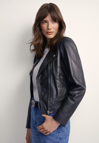 Czarna skórzana kurtka motocyklowa z asymetrycznym zamkiem błyskawicznym, dopasowaną sylwetką i srebrnymi elementami, zestawiona z jasnoszarym topem i niebieskimi dżinsami.