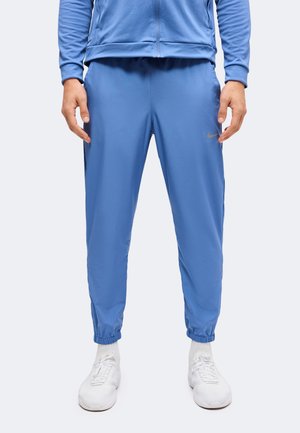 Personne portant un pantalon de jogging Nike bleu, une veste zippée assortie, des chaussettes blanches et des chaussures de sport blanches, debout les mains détendues.