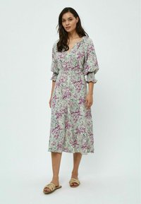 Robe fleurie vert clair avec des motifs violets et roses, présentant un décolleté en V, des manches bouffantes et une longueur midi. Associée à des sandales neutres.