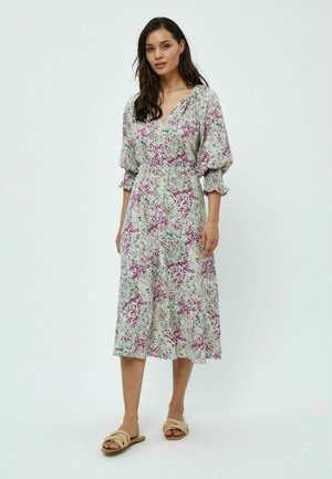 Blumenkleid in hellgrün mit lila und pinken Mustern, mit V-Ausschnitt, Puffärmeln und Midi-Länge. Getragen mit neutralen Sandalen.