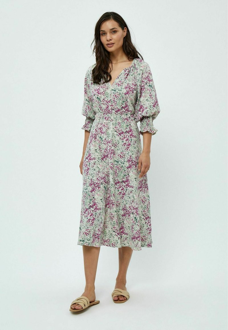 Robe fleurie vert clair avec des motifs violets et roses, présentant un décolleté en V, des manches bouffantes et une longueur midi. Associée à des sandales neutres.