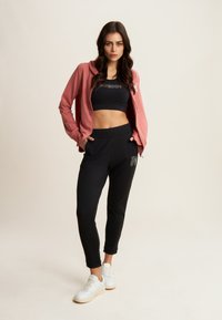 Veste à capuche rose, haut court noir, pantalon jogger noir avec logo, baskets blanches. Le tissu semble doux, coupe décontractée tout au long.