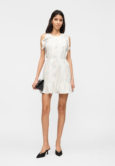 Γυναίκα που φοράει ένα κοντό, λευκό φόρεμα με λουλούδια και βολάν στα μανίκια, κρατώντας μια μαύρη τσάντα χειρός, συνδυασμένο με μαύρες mule γόβες με τακούνι, μπροστά σε απλό φόντο.