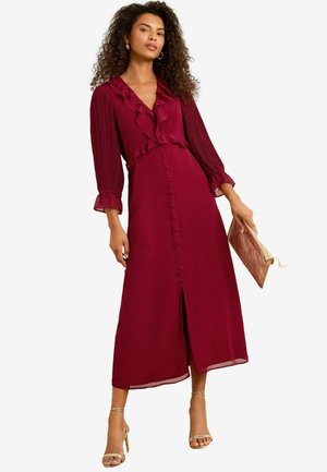 Burgundy midi dress with ruffled neckline and long pleated sleeves. Features a button-down front and a slit. Paired with a textured clutch.