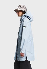 Femme portant un imperméable à capuche bleu clair et un chapeau seau beige avec des taches léopard bleu foncé, debout de profil devant un fond uni.