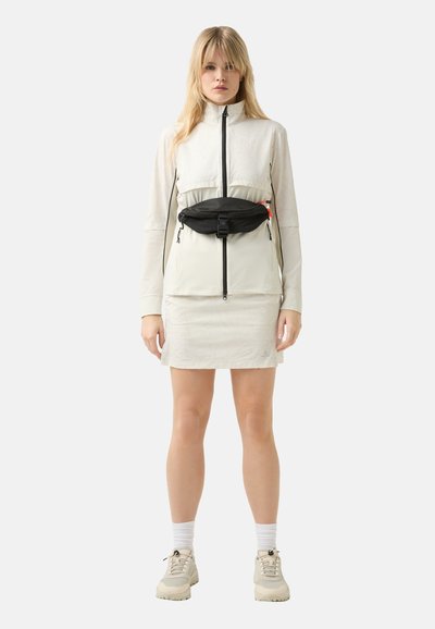 Hellfarbige Jacke mit geometrischem Muster, schwarze Hüfttasche und heller Rock. Die Schuhe sind beige mit schwarzen Akzenten. Glatte, leichte Stoffe.