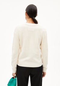 Cremefarbener Pullover mit geripptem Muster, rundem Ausschnitt, Raglanärmeln und einem quadratischen Aufnäher auf der Rückseite. Kombiniert mit schwarzen Hosen.